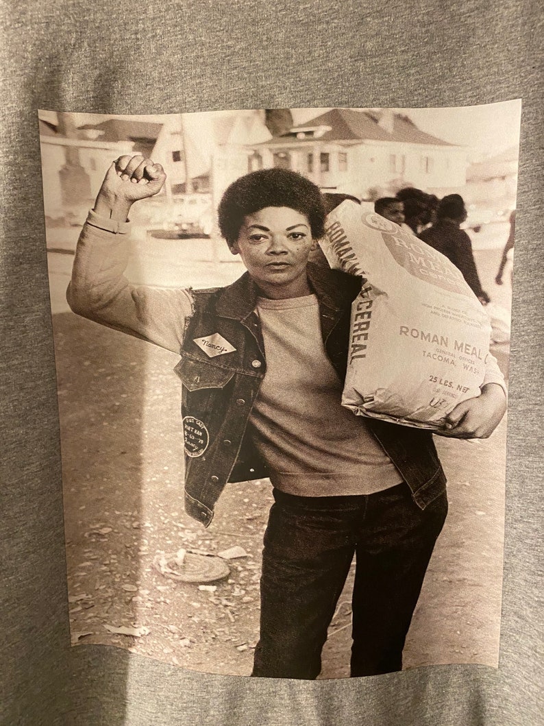 May include: A black and white photograph of a woman raising her fist in the air. She is wearing a denim jacket with a patch that says "Timothy" and is carrying a bag of Roman Meal Cereal. The bag is labeled "Roman Meal Cereal, Enriched, 25 lbs. Net, Tacoma, Wash."
