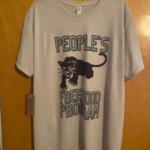 Pode incluir: Uma camiseta cinza claro com um gr&aacute;fico de pantera negra e o texto "PEOPLE'S FREE FOOD PROGRAM" em uma fonte em negrito e com contorno.