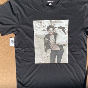 May include: Black t-shirt with a graphic print of a woman holding a bag of flour and raising her fist in the air. The text "K-FUNK 60 XL" is printed on the shirt.