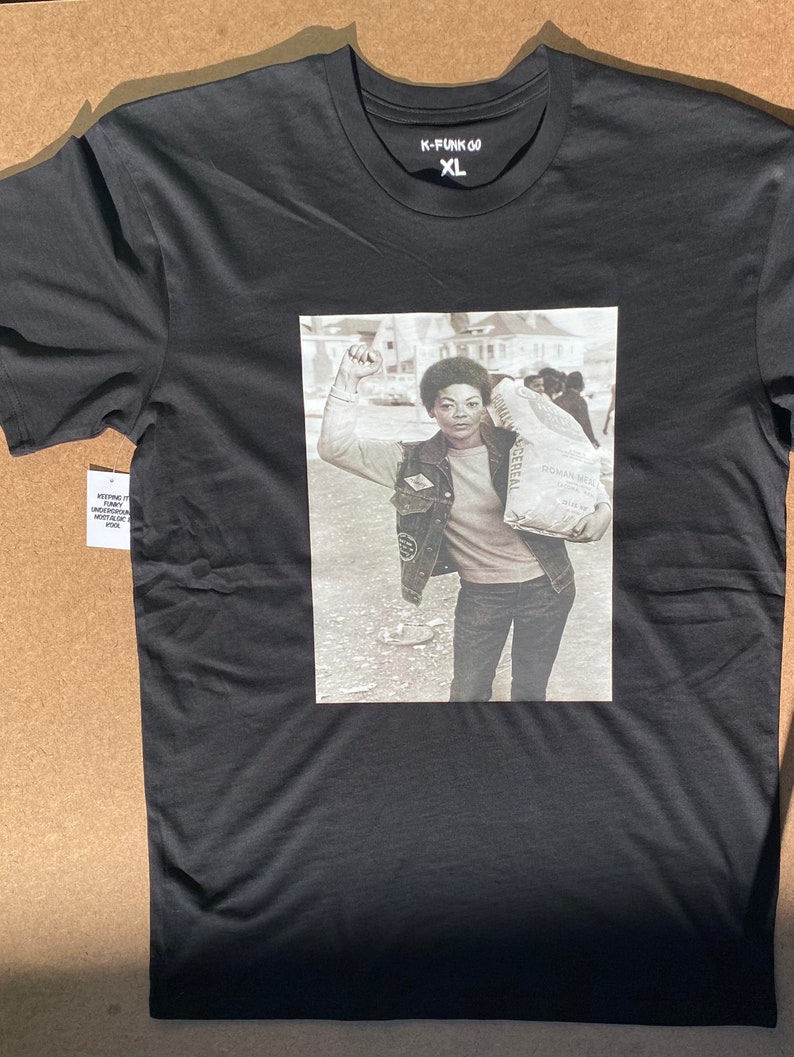 May include: Black t-shirt with a graphic print of a woman holding a bag of flour and raising her fist in the air. The text "K-FUNK 60 XL" is printed on the shirt.