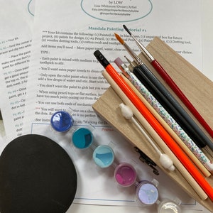May include: A mandala painting tutorial kit with various art supplies. The kit includes paint pots in shades of blue, teal, purple, and white, along with paintbrushes, pencils, cotton swabs, and wooden dotting tools. Instructions are visible in the background.