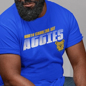 May include: A royal blue t-shirt with a white graphic that says "North Carolina A&T Aggies" in a retro style. A yellow bulldog mascot is featured on the right side of the graphic.