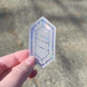 May include: A clear, geometric sticker with a holographic design. The sticker is shaped like a crystal or gem, featuring a detailed outline and internal lines. The design incorporates iridescent elements, creating a shimmering effect.