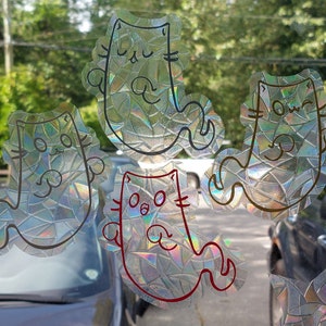 May include: A set of cat-shaped window stickers. The stickers have a holographic, textured finish and are outlined in different colours, including black and red. The cat designs feature simple, cartoonish faces and poses, creating a playful aesthetic.