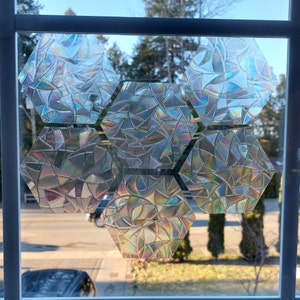 May include: Six iridescent, hexagonal window film panels are attached to a window. The panels have a textured, geometric design that refracts light, creating a rainbow effect. The window frames are white, and the background shows a street scene.
