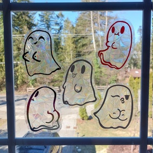 May include: Five holographic ghost window clings with black outlines and different facial expressions. Some ghosts have red accents. The clings are displayed on a window, offering a view of trees and a blue sky in the background.