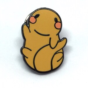 May include: A yellow enamel pin featuring a cartoon character with a smiling face, black outline, and orange cheeks. The pin is in a sitting pose with arms outstretched.
