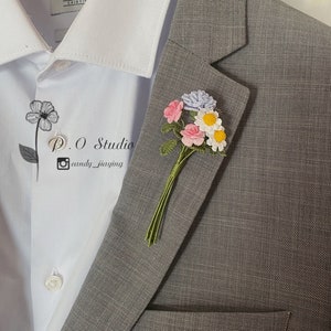 May include: A gray suit jacket with a small bouquet of crocheted flowers pinned to the lapel. The flowers are pink, white, and blue. The text "P.O Studio @candy_jiaying" is visible on the white shirt underneath the jacket.