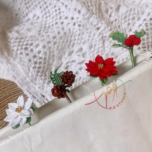 Può includere: Quattro decorazioni di fiori e foglie all'uncinetto, tra cui un fiore bianco, una stella di Natale rossa, una pigna marrone e un rametto di agrifoglio rosso con bacche, sono esposte su uno sfondo di carta bianca con il testo "P.O. STUDIO".