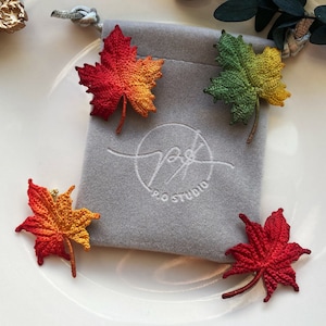 Può includere: Quattro spille a forma di foglia d'acero all'uncinetto in diversi colori: rosso, arancione, giallo e verde. Le spille sono esposte su una pochette in velluto grigio con il testo "P.G. Studio" ricamato su di essa.