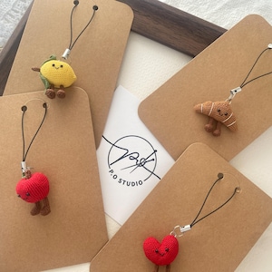 Puede incluir: Encantos de ganchillo hechos a mano con forma de limón, croissant y dos corazones, cada uno con pequeñas patas. Los encantos están unidos a etiquetas de papel marrón con cordones negros. El logotipo de P.O. Studio es visible.