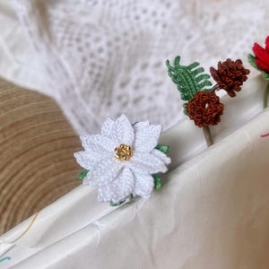 May include: White crocheted poinsettia flower with a gold center, a brown crocheted pine cone, and a green crocheted leaf on a white background.