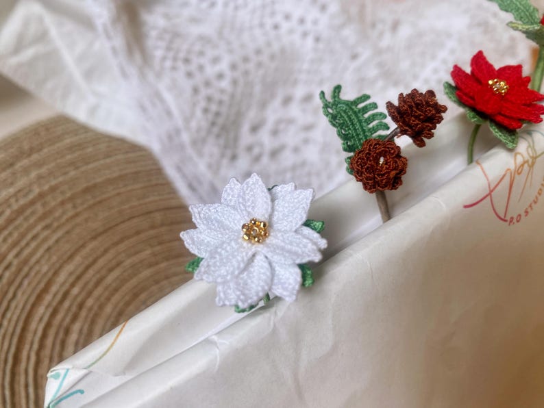 May include: White crocheted poinsettia flower with a gold center, a brown crocheted pine cone, and a green crocheted leaf on a white background.