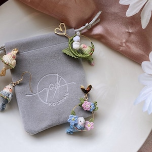 May include: A set of three crocheted earrings featuring small birds and floral designs. The earrings are made with delicate white, blue, green, and pink yarn. The earrings are displayed on a grey surface with a white background.