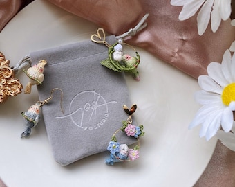 May include: A set of three crocheted earrings featuring small birds and floral designs. The earrings are made with delicate white, blue, green, and pink yarn. The earrings are displayed on a gray surface with a white background.