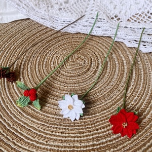 May include: Four crocheted flower pins with green stems on a brown woven surface. The flowers are red, white, and a brown and white combination.