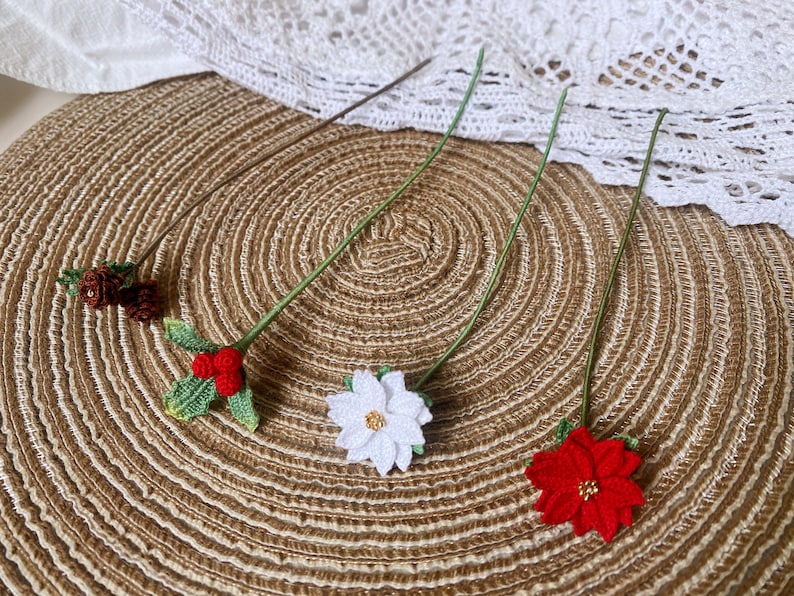 May include: Four crocheted flower pins with green stems on a brown woven surface. The flowers are red, white, and a brown and white combination.