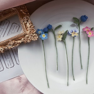 May include: A collection of six crocheted flowers in various colors, including blue, pink, and yellow, displayed on a white plate. The flowers are arranged in a row, with their stems pointing upwards.