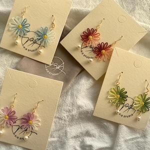 May include: Four pairs of daisy-shaped earrings in various colors: blue, red, green, and purple. Each earring features a gold hook, a delicate floral design, and a small pearl. The earrings are displayed on cream-colored cards with the text "P.O. STUDIO".