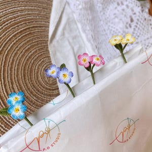 May include: Four crocheted flower bookmarks in different colours: blue, pink, yellow, and white. The bookmarks are arranged on a white paper bag with the text "P.O. Studio" printed on it.