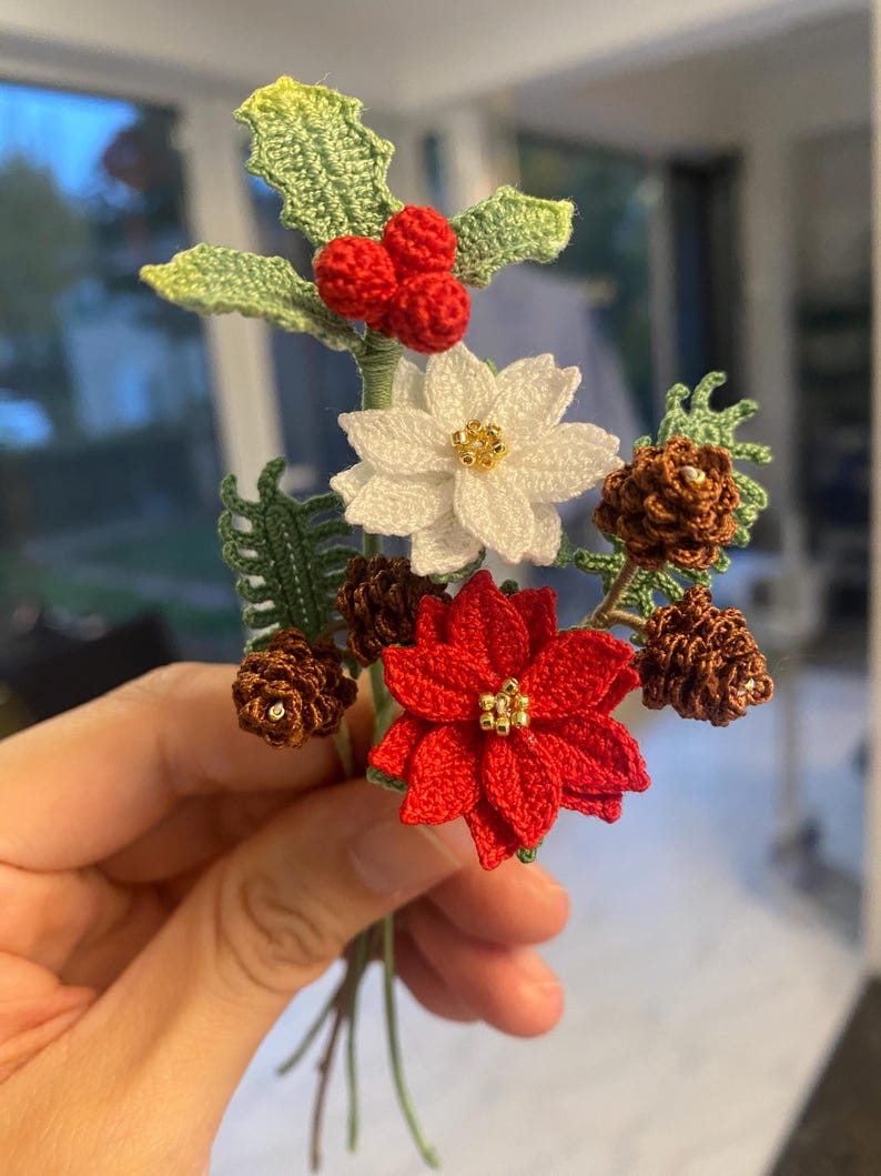 May include: A crocheted Christmas bouquet with red poinsettia, white poinsettia, holly with red berries, and brown pine cones.