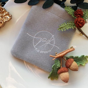 Puede incluir: Un broche de bellota de ganchillo con hojas verdes y un alfiler dorado. El broche está sobre una bolsa de terciopelo gris con el texto "PRO STUDIO" bordado.