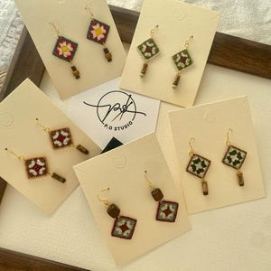 May include: A selection of handmade earrings with square embroidered patterns in red, green, and white. Each earring has a gold-coloured hook and wooden bead detail. The earrings are displayed on cream-coloured cards, presented in a wooden frame.
