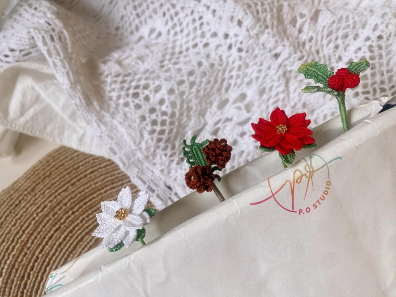 May include: Four crocheted flower and leaf embellishments, including a white flower, a red poinsettia, a brown pine cone, and a red berry holly sprig, are displayed on a white paper background with the text "P.O. STUDIO".