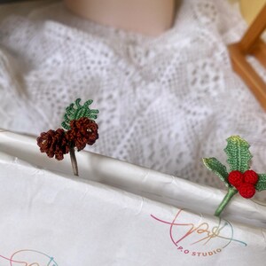 May include: Two crocheted bookmarks, one with a brown pine cone and green leaves, and the other with red berries and green leaves. The bookmarks are on a white surface with the text "P.O. STUDIO" printed on it.