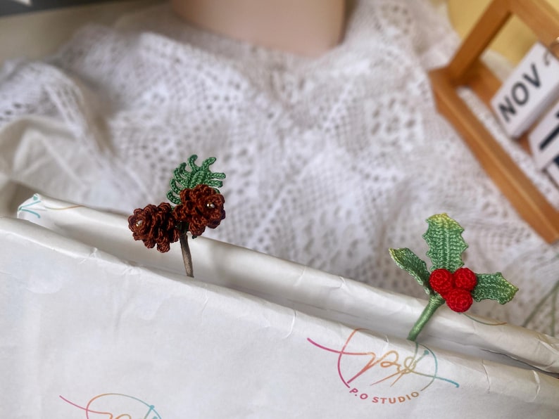 May include: Two crocheted bookmarks, one with a brown pine cone and green leaves, and the other with red berries and green leaves. The bookmarks are on a white surface with the text "P.O. STUDIO" printed on it.
