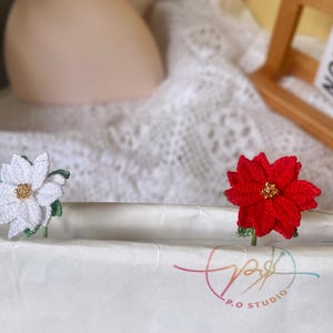 May include: Two crocheted poinsettia flowers, one white and one red, with gold centers, are displayed on a white surface. The flowers are attached to green stems.