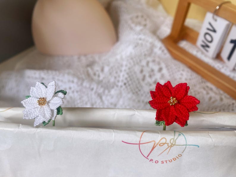 May include: Two crocheted poinsettia flowers, one white and one red, with gold centers, are displayed on a white surface. The flowers are attached to green stems.