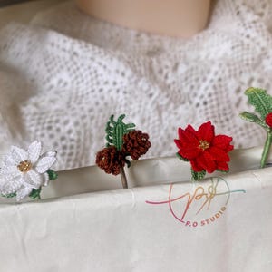May include: Four crocheted Christmas ornaments: a white poinsettia, a brown pine cone, a red poinsettia, and a green holly sprig with red berries. The ornaments are displayed on a white surface with the text "P.O. Studio" visible.