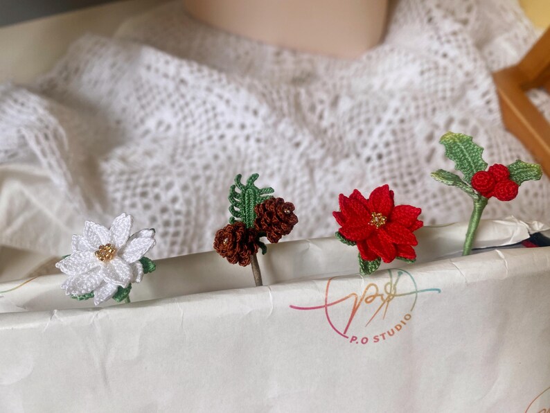 May include: Four crocheted Christmas ornaments: a white poinsettia, a brown pine cone, a red poinsettia, and a green holly sprig with red berries. The ornaments are displayed on a white surface with the text "P.O. Studio" visible.