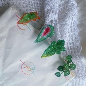 Puede incluir: Cuatro broches de hojas de ganchillo hechos a mano en verde, rojo y naranja. Los broches están sobre un fondo de papel blanco con el texto "P.O. Studio" escrito en él.