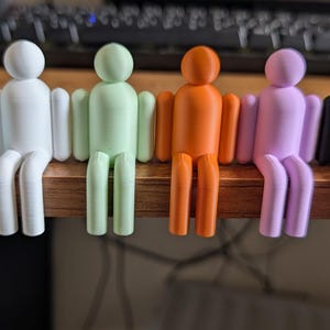 May include: A set of six small, stylised human-shaped figures in a seated position, arranged in a row. The figures are coloured light blue, white, pale green, orange, light purple, and black, each featuring a smooth, rounded head and limbs.