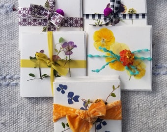 Peut inclure: Une collection de cartes de vœux faites à la main, chacune ornée de fleurs pressées et attachée avec des rubans décoratifs. Les cartes présentent divers arrangements floraux, notamment des fleurs violettes, jaunes et bleues, et sont enveloppées de rubans à motifs.
