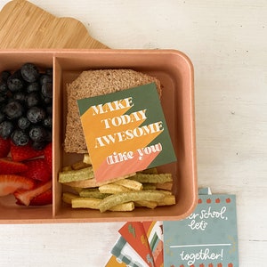 Op de afbeelding: Een roze lunchbox gevuld met blauwe bessen, gesneden aardbeien, een sandwich en groene groentesticks. Een kaart met de tekst "MAKE TODAY AWESOME like you" zit erin. Extra kaarten met schoolthema's zijn ook aanwezig.
