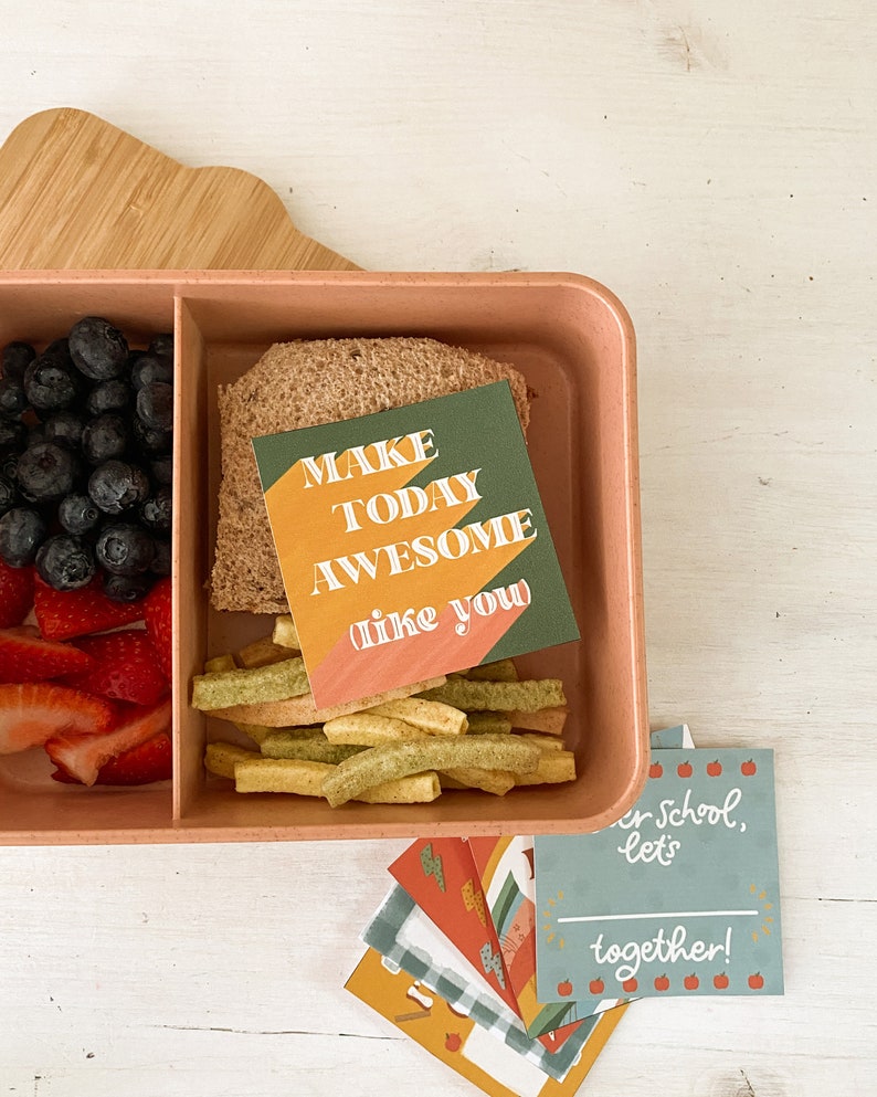 Op de afbeelding: Een roze lunchbox gevuld met blauwe bessen, gesneden aardbeien, een sandwich en groene groentesticks. Een kaart met de tekst "MAKE TODAY AWESOME like you" zit erin. Extra kaarten met schoolthema's zijn ook aanwezig.