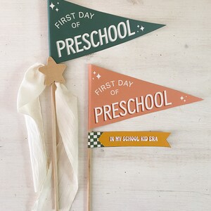 Op de afbeelding: Houten wimpelvlaggen voor de eerste schooldag van de kleuterschool. De vlaggen zijn groen en perzik met witte tekst die luidt "First Day of Preschool". Een kleinere gele vlag luidt "In My School Kid Era".