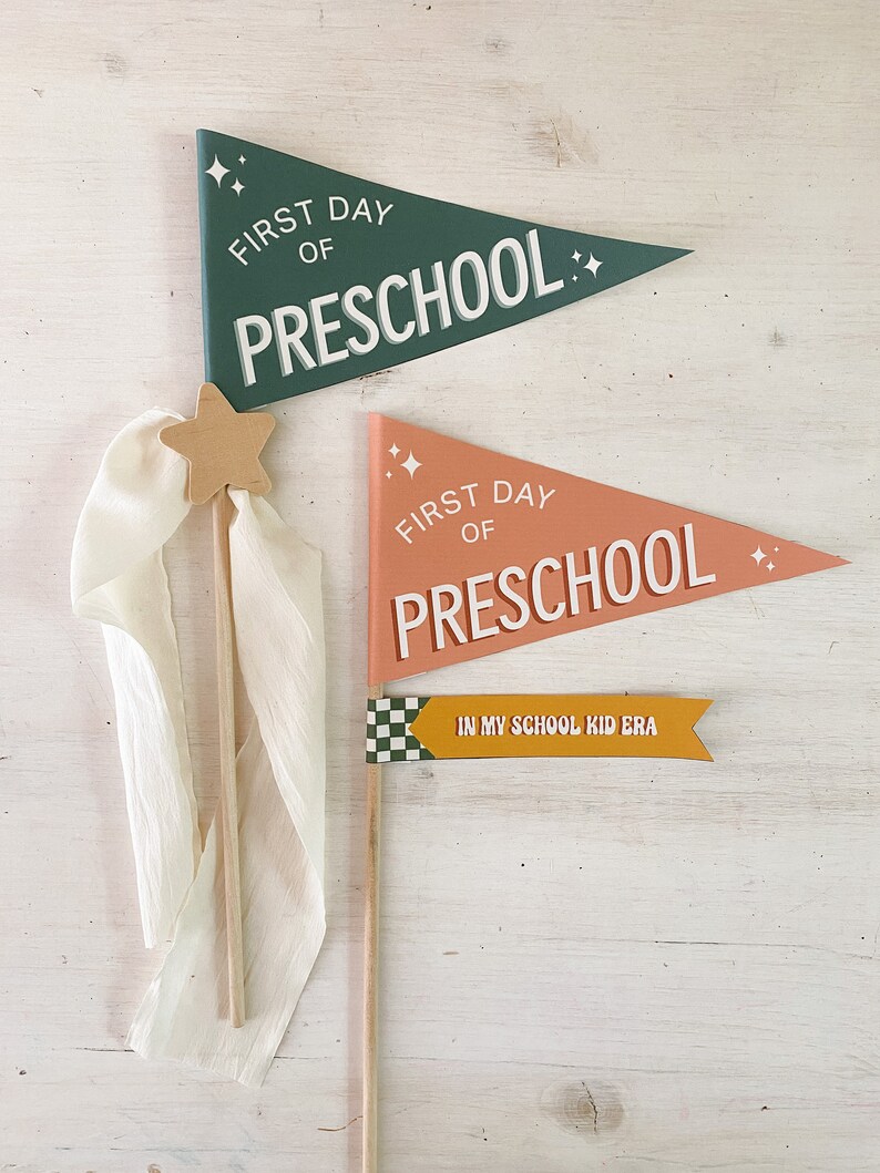 Op de afbeelding: Houten wimpelvlaggen voor de eerste schooldag van de kleuterschool. De vlaggen zijn groen en perzik met witte tekst die luidt "First Day of Preschool". Een kleinere gele vlag luidt "In My School Kid Era".