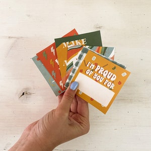 Op de afbeelding: Een hand die een waaier van kleurrijke wenskaarten vasthoudt. De kaarten hebben verschillende ontwerpen, waaronder bliksemschichten, ruitpatronen en de tekst "I'M PROUD OF YOU FOR". De kaarten zijn in tinten oranje, groen en blauw.