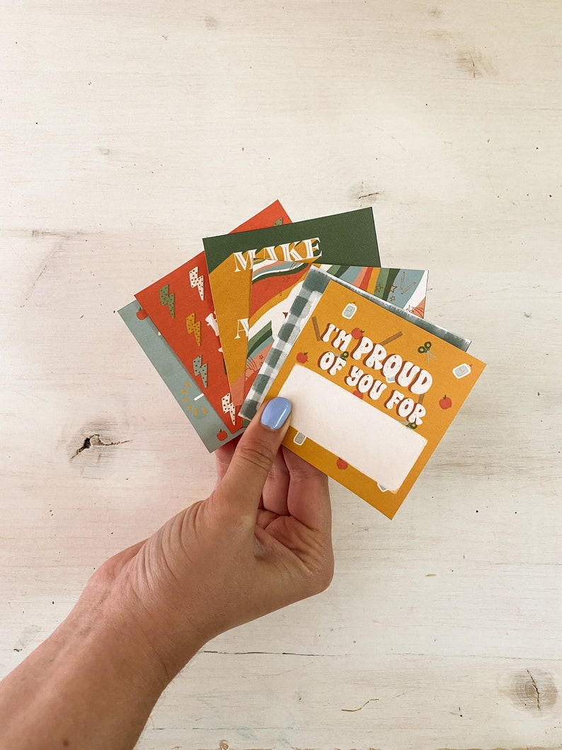 Op de afbeelding: Een hand die een waaier van kleurrijke wenskaarten vasthoudt. De kaarten hebben verschillende ontwerpen, waaronder bliksemschichten, ruitpatronen en de tekst "I'M PROUD OF YOU FOR". De kaarten zijn in tinten oranje, groen en blauw.