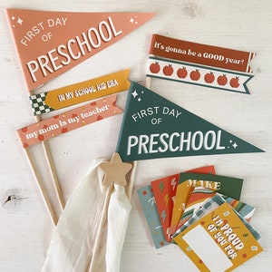 Op de afbeelding: Een verzameling schooldecoraties, waaronder wimpels en kleine kaarten. De vlaggen tonen zinnen als "First Day of Preschool" en "It's gonna be a GOOD year!" in witte letters. Het kleurenpalet omvat perzik, groen en geel.