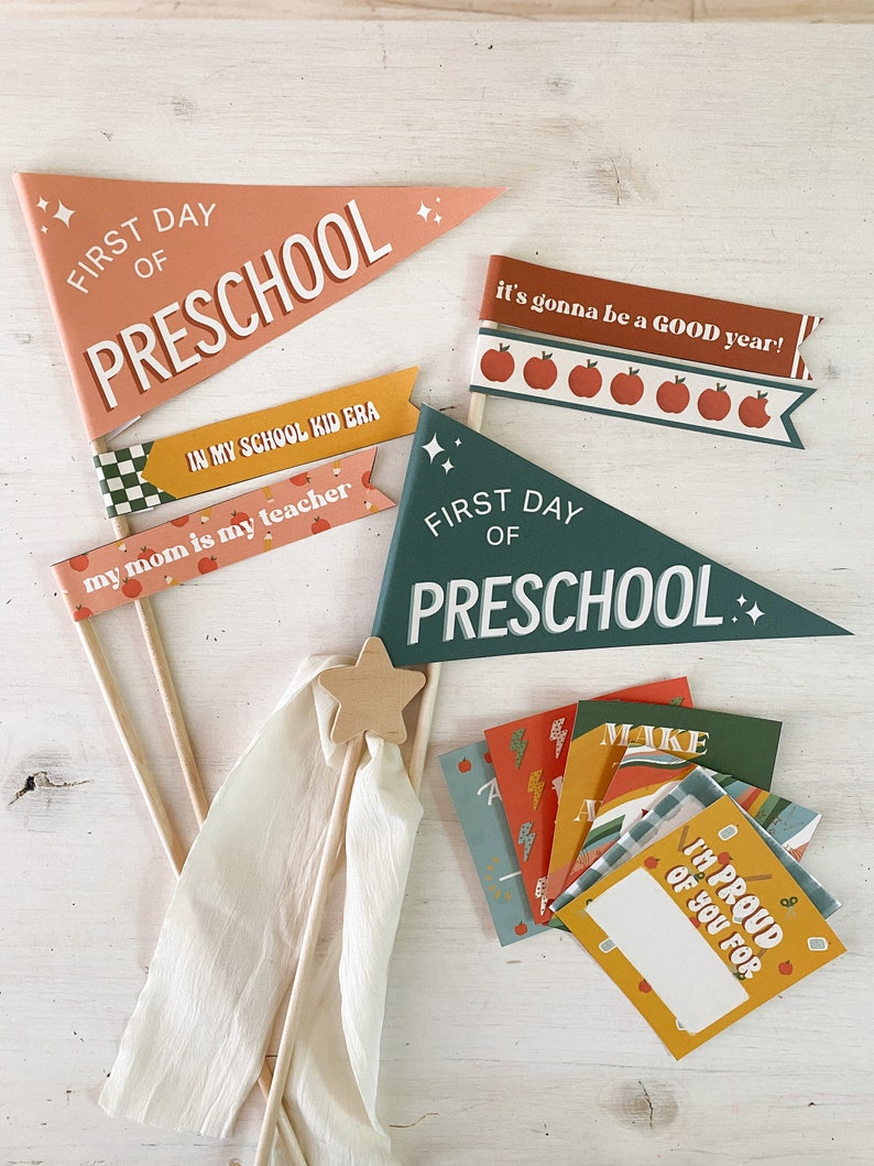 Op de afbeelding: Een verzameling schooldecoraties, waaronder wimpels en kleine kaarten. De vlaggen tonen zinnen als "First Day of Preschool" en "It's gonna be a GOOD year!" in witte letters. Het kleurenpalet omvat perzik, groen en geel.