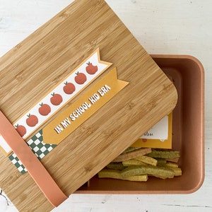 Op de afbeelding: Een bamboe lunchbox met een lichtbruin deksel en een perzikkleurige container. Het deksel heeft decoratieve banners met appel illustraties en de tekst "IN MY SCHOOL KID ERA". De container bevat kleurrijke snacks.