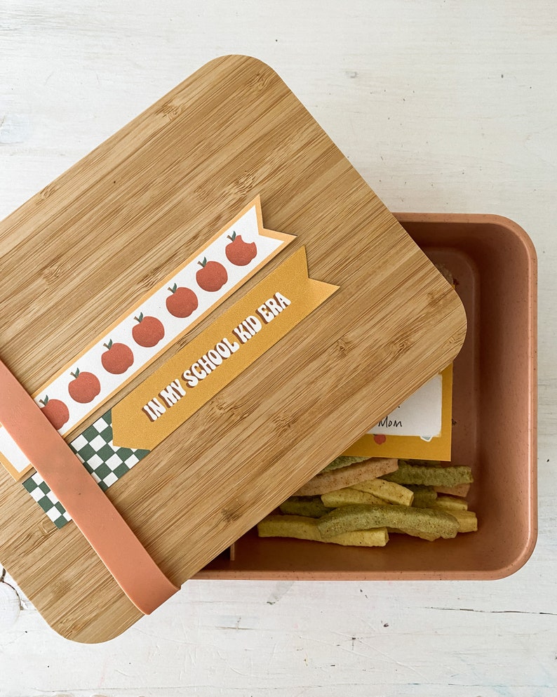 Op de afbeelding: Een bamboe lunchbox met een lichtbruin deksel en een perzikkleurige container. Het deksel heeft decoratieve banners met appel illustraties en de tekst "IN MY SCHOOL KID ERA". De container bevat kleurrijke snacks.