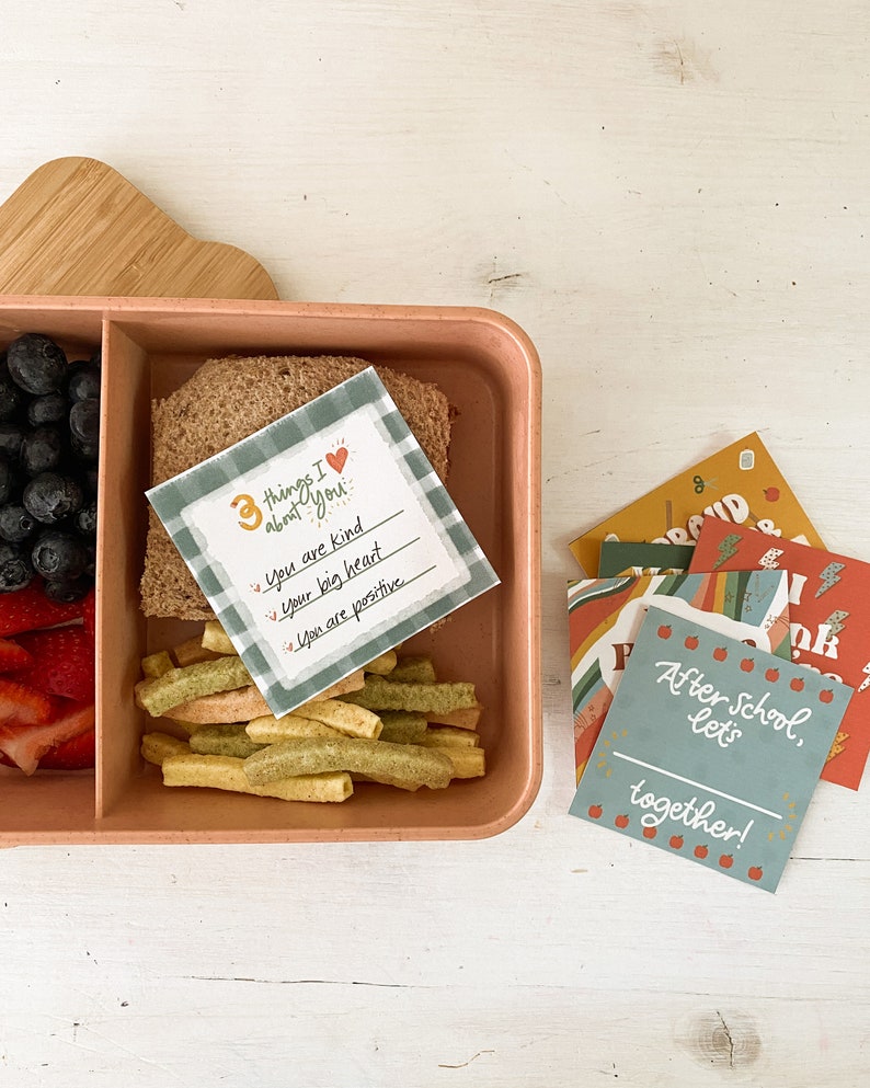 Op de afbeelding: Een perzikkleurige lunchbox met compartimenten, gevuld met blauwe bessen, aardbeien, een sandwich en groene en gele snacks. Er is ook een kaart met de tekst "3 things I about you" en andere kaarten met positieve boodschappen.
