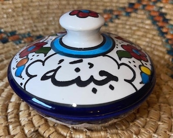 Könnte beinhalten: Eine weiße Keramikschale mit einem blauen und weißen Blumenmuster. Die Schale hat einen Deckel mit einem Knopf und das Wort "جنية" in arabischer Schrift.