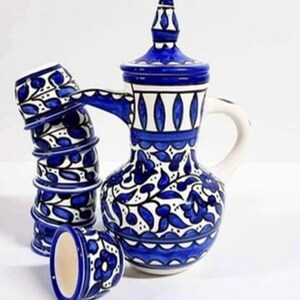 Puede incluir: Una tetera de cerámica azul y blanca con un juego a juego de seis tazas. La tetera tiene un diseño floral y una tapa con una parte superior puntiaguda. Las tazas están apiladas una encima de la otra.